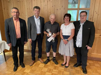 Fünf Erwachsene stehen in einem Raum mit Holzwänden und lächeln für ein Foto. Der Mann in der Mitte hält ein eingewickeltes Geschenk und eine Flasche. Die Frau neben ihm trägt eine weiße Bluse und einen Rock. Hinter ihnen spiegelt ein Glasfenster die Außenwelt wider.