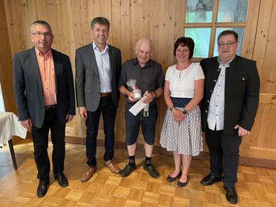 Fünf Erwachsene stehen in einem Raum mit Holzwänden und lächeln für ein Foto. Der Mann in der Mitte hält ein eingewickeltes Geschenk und eine Flasche. Die Frau neben ihm trägt eine weiße Bluse und einen Rock. Hinter ihnen spiegelt ein Glasfenster die Außenwelt wider.