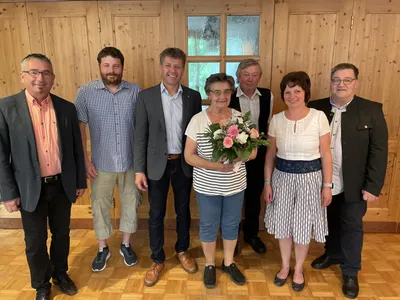 Eine Gruppe von sechs Personen, darunter zwei Frauen, steht vor einer Holzwand mit einem Fenster. Die Frau in der Mitte hält einen Blumenstrauß.