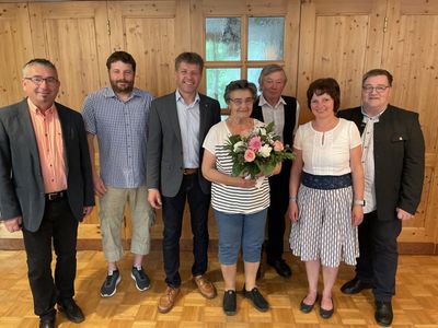 Eine Gruppe von sechs Personen, darunter zwei Frauen, steht vor einer Holzwand mit einem Fenster. Die Frau in der Mitte hält einen Blumenstrauß.