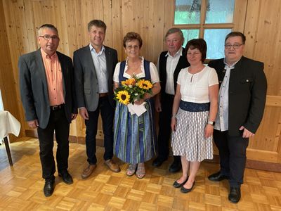 Fünf Personen, darunter eine Frau mit einem Strauß Sonnenblumen, stehen in einem Raum mit Holzwänden und einem Holzboden.