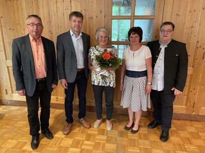Fünf Menschen stehen in einem Raum mit Holzwänden und einem Glasfenster. Die Frau in der Mitte hält einen Blumenstrauß. Der Mann auf der linken Seite trägt einen Gürtel und eine Brille. Der Mann auf der rechten Seite trägt einen schwarzen Anzug.