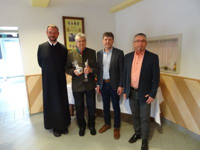 Fünf Männer stehen in einem Raum, einer hält eine Trophäe und eine Flasche, während ein anderer einen schwarzen Umhang trägt. Hinter ihnen steht ein Tisch mit einer weißen Tischdecke.