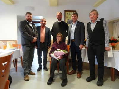 Eine Gruppe von sechs Personen, darunter eine ältere Frau mit Blumen, steht in einem Raum mit weißer Wand und gefliestem Boden.
