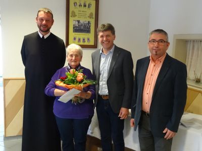 Vier Personen stehen lächelnd für ein Foto zusammen. Eine Frau hält einen Blumenstrauß. Hinter ihnen hängt ein gerahmtes Bild an der Wand.