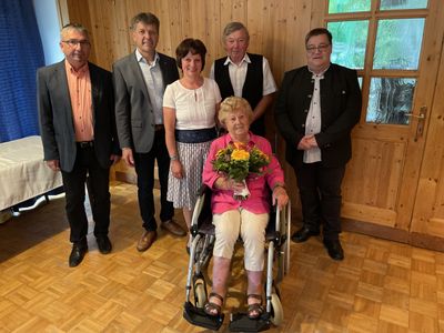 Eine Gruppe von Menschen steht und sitzt in einem Raum, wobei eine Frau im Rollstuhl einen Blumenstrauß hält.
