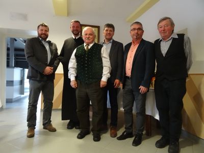 Sechs Männer in formeller Kleidung, einschließlich eines Priesters, stehen zusammen in einem Raum.