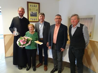 Fünf Personen stehen zusammen in einem Raum, eine Frau hält Blumen und ein Mann in einem schwarzen Gewand hält ein kleines Geschenk. Hinter ihnen ist eine weiße Wand mit einem gerahmten Foto.