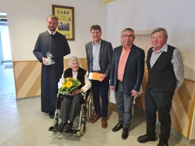 Eine Gruppe von Menschen, einschließlich eines Priesters, steht in einem Raum mit einer Frau im Rollstuhl, die Blumen hält.