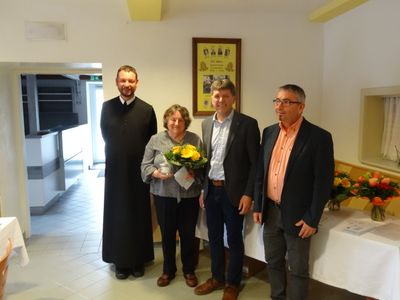 Vier Menschen stehen in einem Raum und lächeln. Eine Frau hält einen Blumenstrauß. Dahinter ist ein gerahmtes Foto an der Wand befestigt.