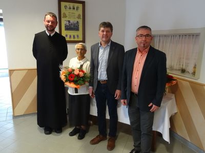 Vier Personen, darunter eine Frau mit Blumen, stehen in einem Raum mit einem Tisch und einem gerahmten Bild an der Wand.