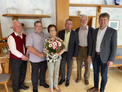 Eine Gruppe von sechs Erwachsenen, darunter eine Frau mit einem Blumenstrauß, steht in einem Raum mit Holzböden und Regalen.