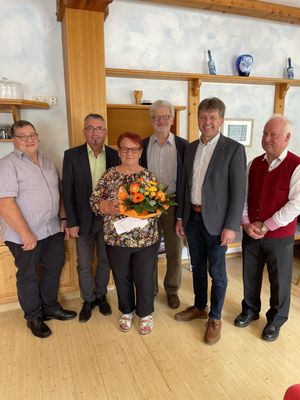 Eine Gruppe von sechs Personen steht in einem Raum und lächelt für ein Foto. Eine Frau in der Mitte hält einen Blumenstrauß. Dahinter befindet sich ein Holzregal mit verschiedenen Gegenständen.