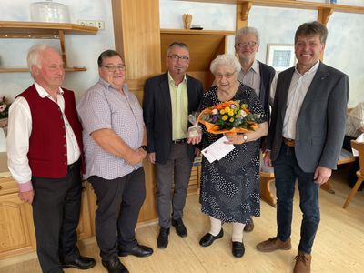 Eine Gruppe von sechs Personen, darunter eine ältere Frau mit Blumen und einem Zertifikat, steht in einem Raum mit Holzböden und Regalen.