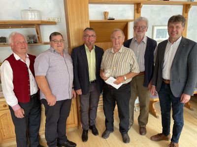 Sechs Männer stehen in einem Raum, einer hält eine Flasche und Papier. Sie tragen Freizeitkleidung und scheinen für ein Foto zu posieren. Hinter ihnen sind ein Holzschrank und Regale zu sehen.