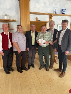 Sechs Männer stehen in einem Raum, lächeln und posieren für ein Foto. Der Mann in der Mitte hält eine Flasche und Papier. Hinter ihnen sind hölzerne Regale mit verschiedenen Gegenständen zu sehen.