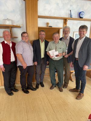 Sechs Männer stehen in einem Raum, lächeln und posieren für ein Foto. Der Mann in der Mitte hält eine Flasche und Papier. Hinter ihnen sind hölzerne Regale mit verschiedenen Gegenständen zu sehen.