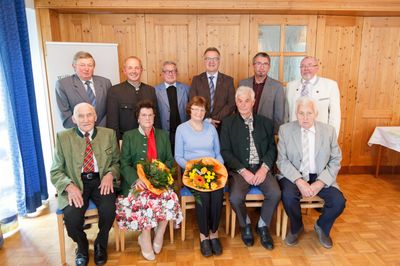Eine Gruppe älterer Personen, sowohl Männer als auch Frauen, sitzt in einem Raum mit Holzwänden und -böden, gekleidet in formeller Kleidung und hält Blumen.