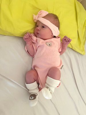 Ein Babymädchen in rosa Kleidung mit einem rosa Stirnband und Socken mit Bärenabdrücken liegt auf einem Bett mit einem gelben Kissen.
