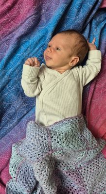 Ein Baby liegt auf einer bunten Decke und trägt einen cremefarbenen Strickpullover mit V-Ausschnitt, es scheint zu lächeln.
