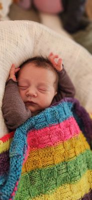 Ein Neugeborenes schläft friedlich auf einer weißen Decke, eingewickelt in eine bunte gestrickte Decke. Das Baby trägt ein braunes Langarmshirt.
