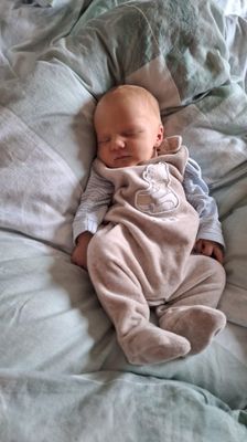 Ein Baby schläft friedlich auf einem Bett, trägt ein blau-weiß gestreiftes Langarmshirt und einen beigen Overall mit einem Cartoon-Charakter.