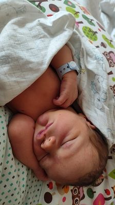 Ein Neugeborenes schläft auf einem Bett, das mit einer weißen Decke und einer bunten Decke mit Cartoon-Tieren bedeckt ist. Das Baby trägt ein blaues Armband.