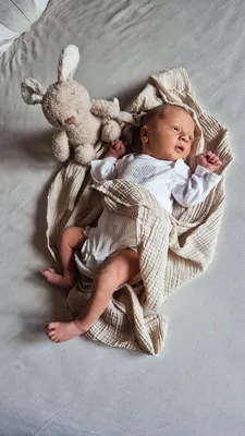 Ein Baby liegt auf einem Bett, eingewickelt in eine beige Decke, mit einem weißen Plüschkaninchen neben ihm. Das Baby ist in Weiß gekleidet und scheint zu schlafen.