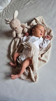 Ein Baby liegt auf einem Bett, eingewickelt in eine beige Decke, mit einem weißen Plüschkaninchen neben ihm. Das Baby ist in Weiß gekleidet und scheint zu schlafen.