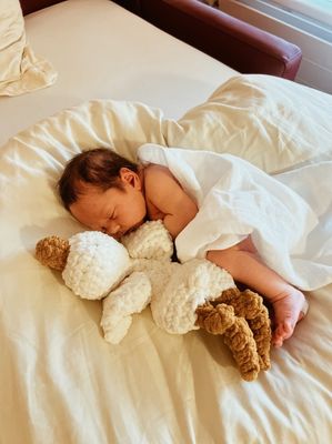 Ein Baby schläft friedlich auf einem Bett, eingewickelt in eine weiße Decke, mit einem Kuscheltier neben ihm.
