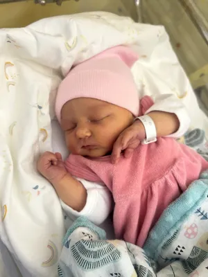 Ein neugeborenes Mädchen schläft friedlich in einem Krankenhausbett, trägt eine rosa Mütze und ein rosa Kleid. Sie hat ein weißes Krankenhausarmband am Handgelenk.