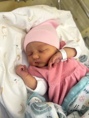 Ein neugeborenes Mädchen schläft friedlich in einem Krankenhausbett, trägt eine rosa Mütze und ein rosa Kleid. Sie hat ein weißes Krankenhausarmband am Handgelenk.