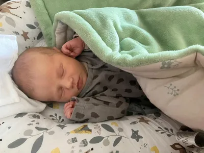 Ein Baby schläft friedlich unter einer mintgrünen Decke und trägt ein graues Strampler mit braunen Flecken. Der Kopf des Babys ruht auf einem Kissen mit Blattmuster.