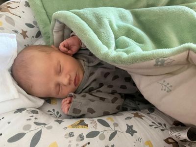 Ein Baby schläft friedlich unter einer mintgrünen Decke und trägt ein graues Strampler mit braunen Flecken. Der Kopf des Babys ruht auf einem Kissen mit Blattmuster.