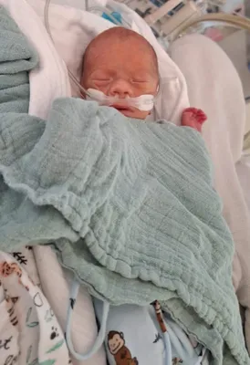 Ein Neugeborenes liegt auf einem Bett, eingewickelt in eine hellblaue Decke, mit einem Sauerstoffschlauch an der Nase. Die Augen des Babys sind geschlossen, und es scheint friedlich zu schlafen.