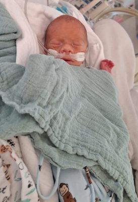 Ein Neugeborenes liegt auf einem Bett, eingewickelt in eine hellblaue Decke, mit einem Sauerstoffschlauch an der Nase. Die Augen des Babys sind geschlossen, und es scheint friedlich zu schlafen.