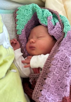 Ein Neugeborenes schläft friedlich eingewickelt in eine bunte gehäkelte Decke. Das Gesicht des Babys zeigt einen ruhigen Ausdruck mit geschlossenen Augen.