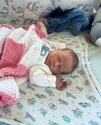 Ein Baby schläft friedlich auf einem Bett, das mit einer pink-weißen gehäkelten Decke bedeckt ist, mit einem blauen Plüschtier in der Nähe.