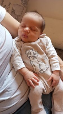 Ein schlafendes Baby in einem Simba-Themen-Overall, das von einem Erwachsenen auf einer Couch gehalten wird.