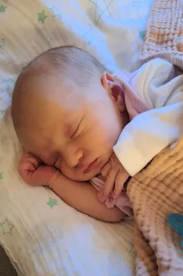 Ein Babymädchen schläft friedlich auf einem Bett mit einer rosa Decke und einer weißen Decke. Sie trägt ein rosa Outfit und hat ihre Hand im Gesicht.