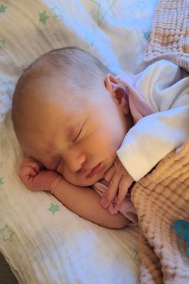 Ein Babymädchen schläft friedlich auf einem Bett mit einer rosa Decke und einer weißen Decke. Sie trägt ein rosa Outfit und hat ihre Hand im Gesicht.