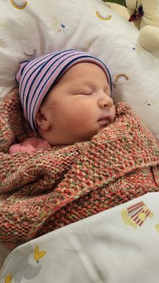 Ein Baby schläft friedlich, eingewickelt in eine gestrickte Decke, mit einer gestreiften Mütze. Das Gesicht des Babys ist teilweise sichtbar und zeigt einen ruhigen Ausdruck.