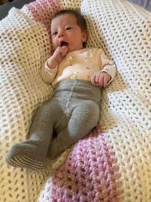 Ein Baby liegt auf einer gestrickten Decke und trägt ein beiges Langarmshirt mit Hasendesign und graue Hose. Das Baby schaut mit der Hand in der Nähe des Mundes nach oben.