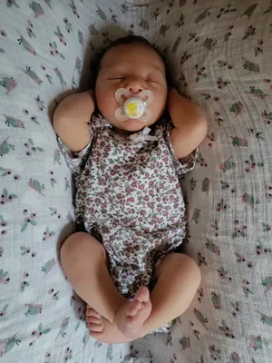 Ein Babymädchen schläft auf einer floral gemusterten Decke, trägt ein Blumenbody und einen Schnuller mit einem gelben Bienendesign.