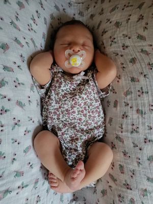 Ein Babymädchen schläft auf einer floral gemusterten Decke, trägt ein Blumenbody und einen Schnuller mit einem gelben Bienendesign.