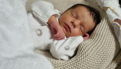 Ein Neugeborenes schläft friedlich auf einer gestrickten Decke und trägt ein weißes Strampler mit Igel-Design. Die Hand des Babys liegt sanft auf der Wange.