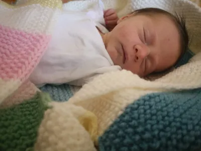 Ein neugeborenes Baby schläft friedlich auf einer bunten gehäkelten Decke, mit einer Hand auf der Wange.