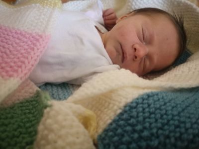 Ein neugeborenes Baby schläft friedlich auf einer bunten gehäkelten Decke, mit einer Hand auf der Wange.
