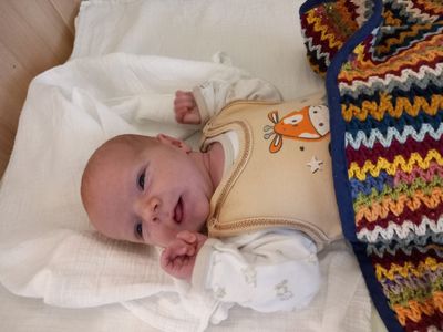Ein Baby liegt auf einer weißen Decke und trägt ein beiges Lätzchen mit einem Giraffendesign. Die Hände des Babys sind erhoben, und es scheint zu lächeln.