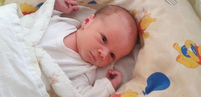 Ein Nahaufnahme eines Babys, das auf einem Bett mit einer weißen Decke liegt, ein weißes Shirt trägt und lächelt.